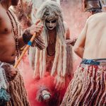 Participantes indígenas dançam em uma apresentação cultural ao ar livre, usando trajes tradicionais e adornos coloridos.