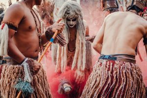 Participantes indígenas dançam em uma apresentação cultural ao ar livre, usando trajes tradicionais e adornos coloridos.