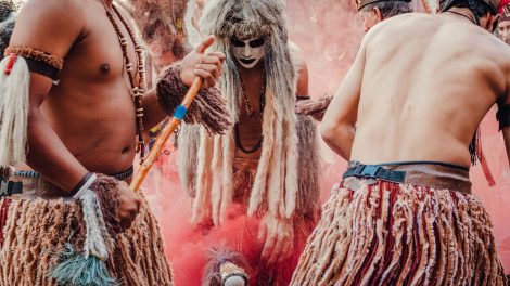 Participantes indígenas dançam em uma apresentação cultural ao ar livre, usando trajes tradicionais e adornos coloridos.