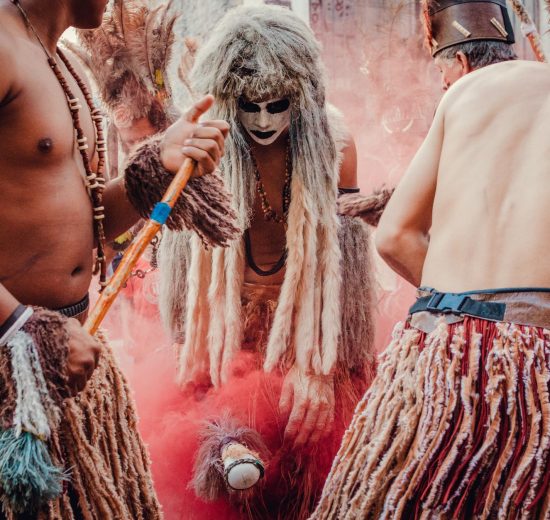 Participantes indígenas dançam em uma apresentação cultural ao ar livre, usando trajes tradicionais e adornos coloridos.