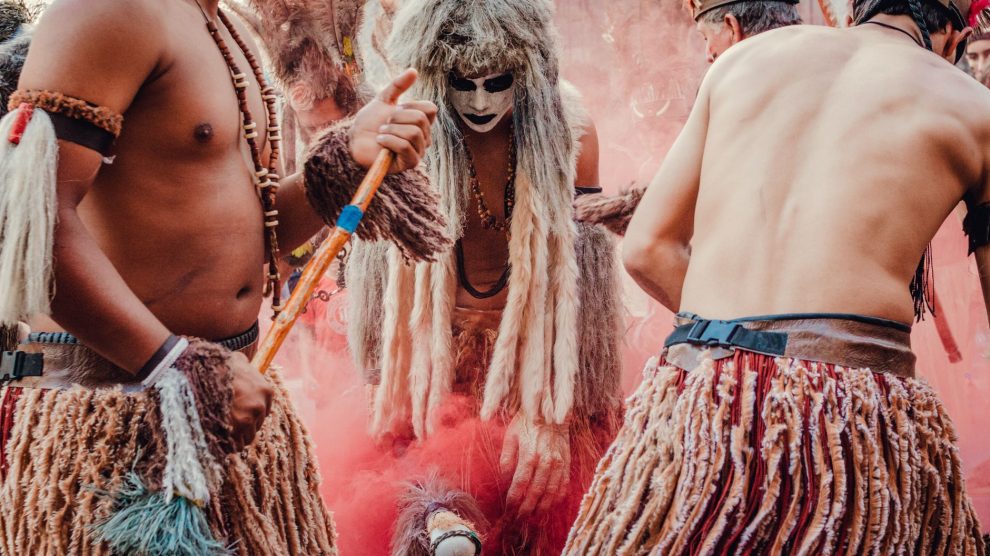 Participantes indígenas dançam em uma apresentação cultural ao ar livre, usando trajes tradicionais e adornos coloridos.