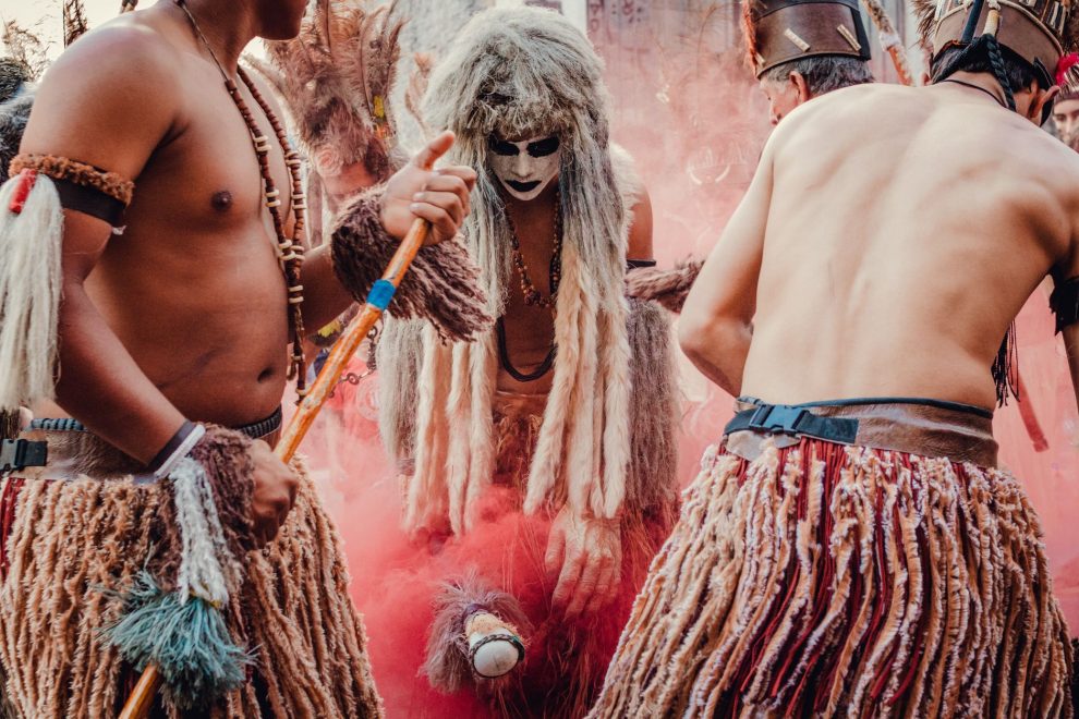 Participantes indígenas dançam em uma apresentação cultural ao ar livre, usando trajes tradicionais e adornos coloridos.