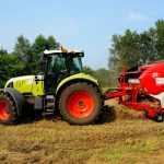 Trator verde equipado com enfardadora trabalhando em um campo agrícola, com árvores ao fundo.