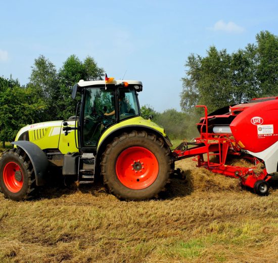 Trator verde equipado com enfardadora trabalhando em um campo agrícola, com árvores ao fundo.