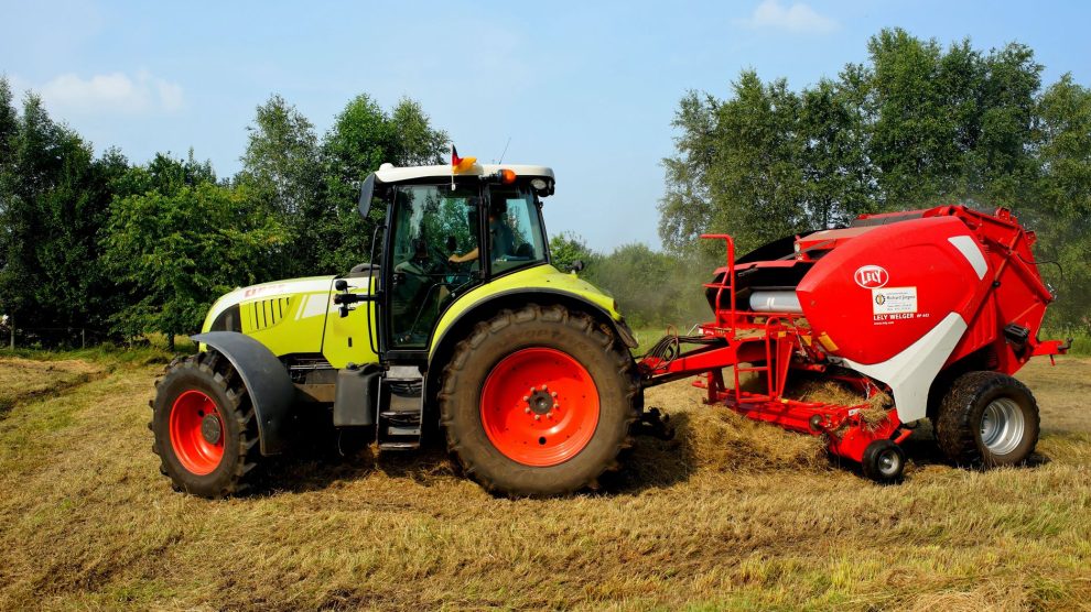 Trator verde equipado com enfardadora trabalhando em um campo agrícola, com árvores ao fundo.