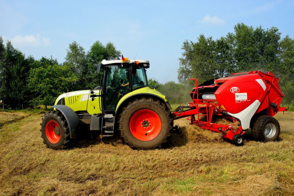 Trator verde equipado com enfardadora trabalhando em um campo agrícola, com árvores ao fundo.
