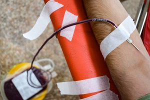 Braço de uma pessoa durante doação de sangue, com agulha inserida e bolsa coletora visível.