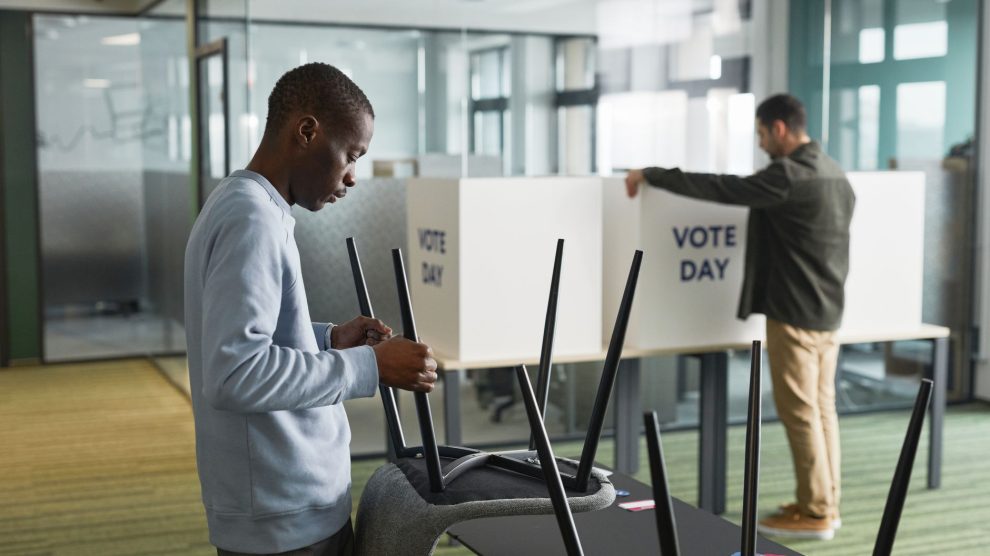 Homens preparam cabines de votação em um ambiente interno para o dia da eleição.