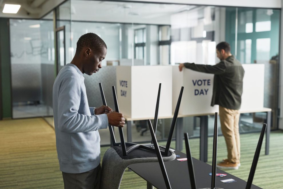 Homens preparam cabines de votação em um ambiente interno para o dia da eleição.
