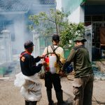 Três agentes realizam fumigação em uma rua residencial para combater mosquitos.