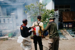 Três agentes realizam fumigação em uma rua residencial para combater mosquitos.