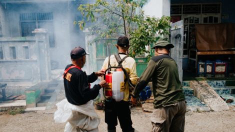 Três agentes realizam fumigação em uma rua residencial para combater mosquitos.