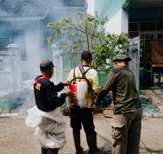 Três agentes realizam fumigação em uma rua residencial para combater mosquitos.