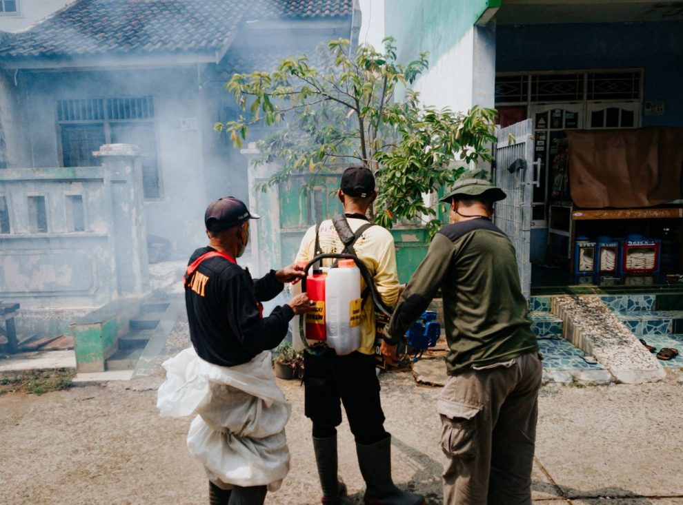 Três agentes realizam fumigação em uma rua residencial para combater mosquitos.
