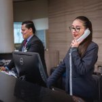 Profissionais trabalham em um balcão de atendimento, com uma mulher ao telefone e um homem ao lado.