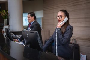 Profissionais trabalham em um balcão de atendimento, com uma mulher ao telefone e um homem ao lado.
